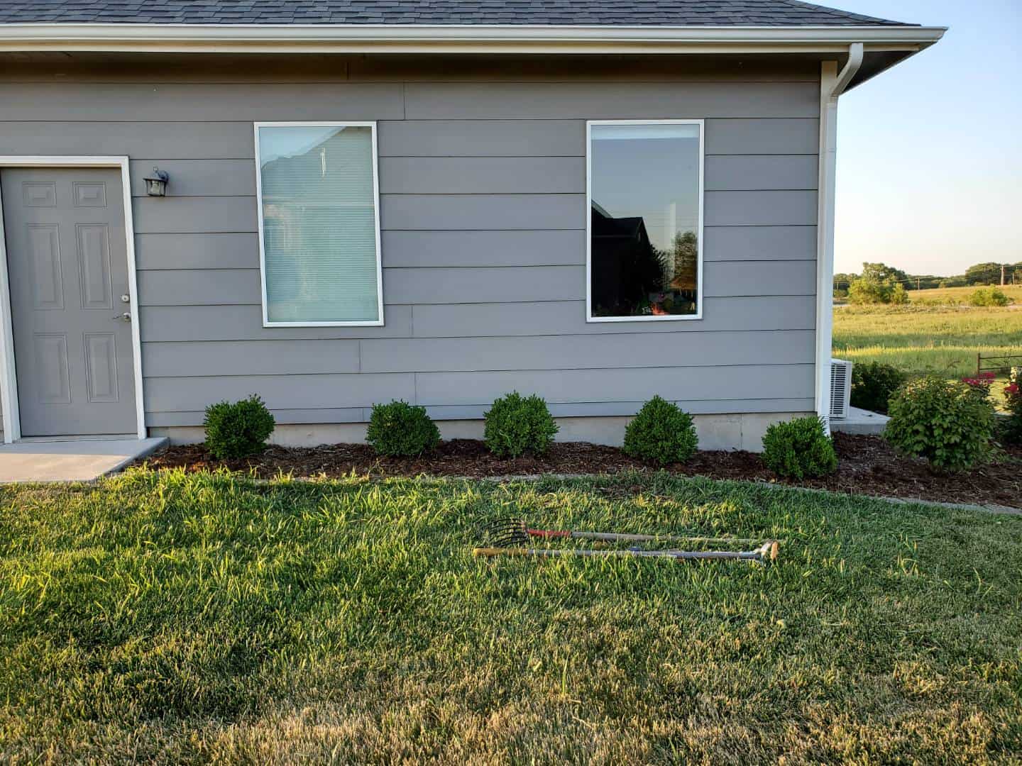 Freshly edged flower bed with evenly spaced shrubs in front of a gray house, with garden tools resting on the lawn in the foreground.