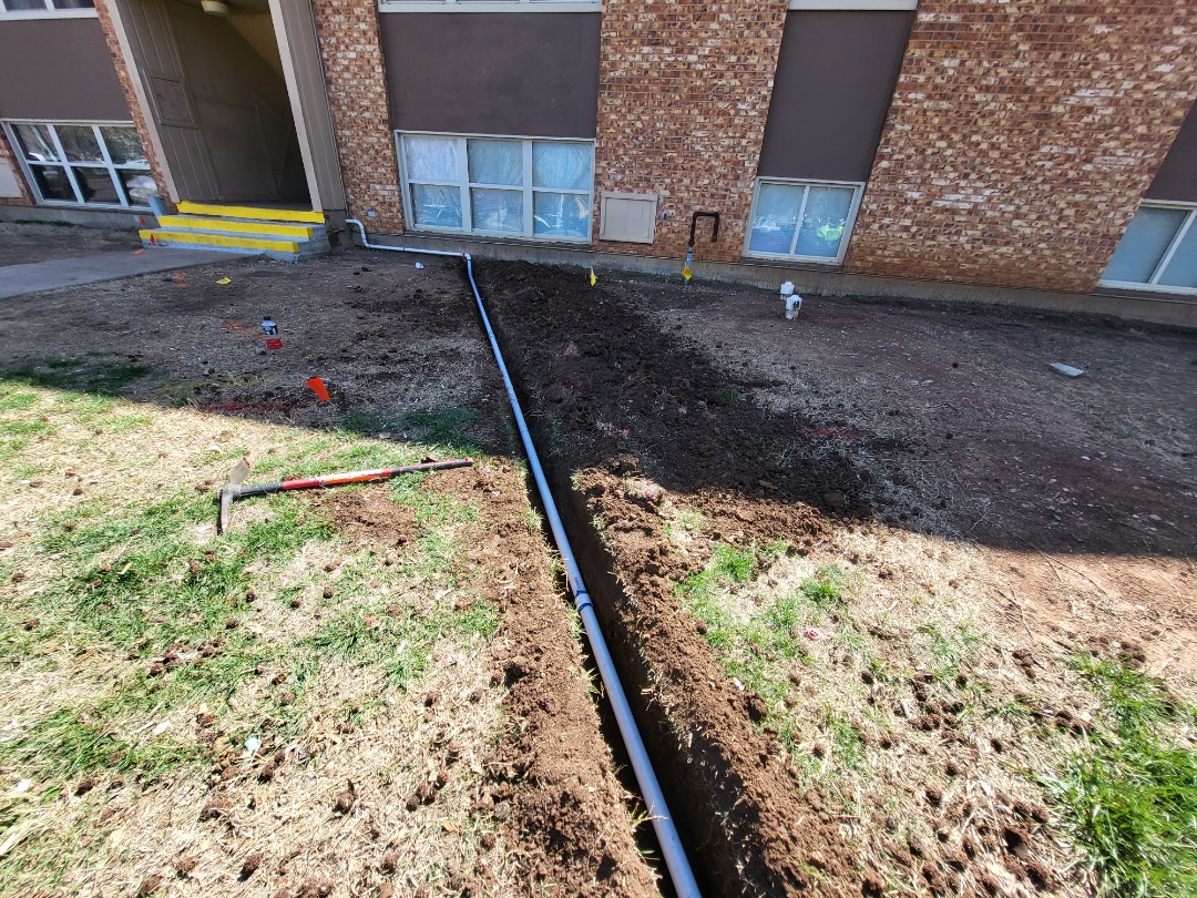 New drainage pipe installed in a trench beside a brick building with yellow stair steps and landscaping tools visible on the ground.