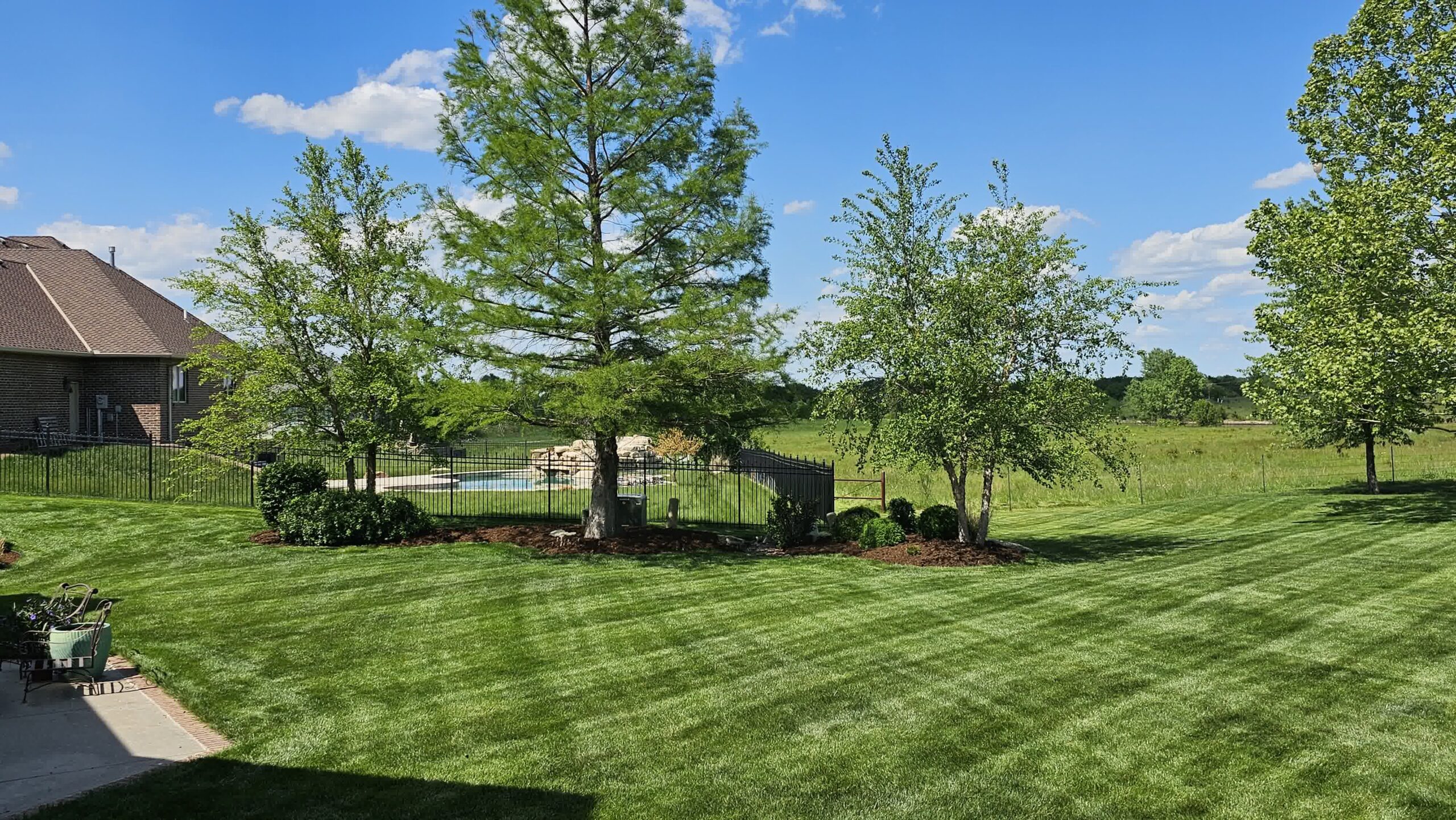 Spacious backyard with striped green lawn, trees, mulch beds, and black iron fence enclosing an in-ground pool and waterfall feature.