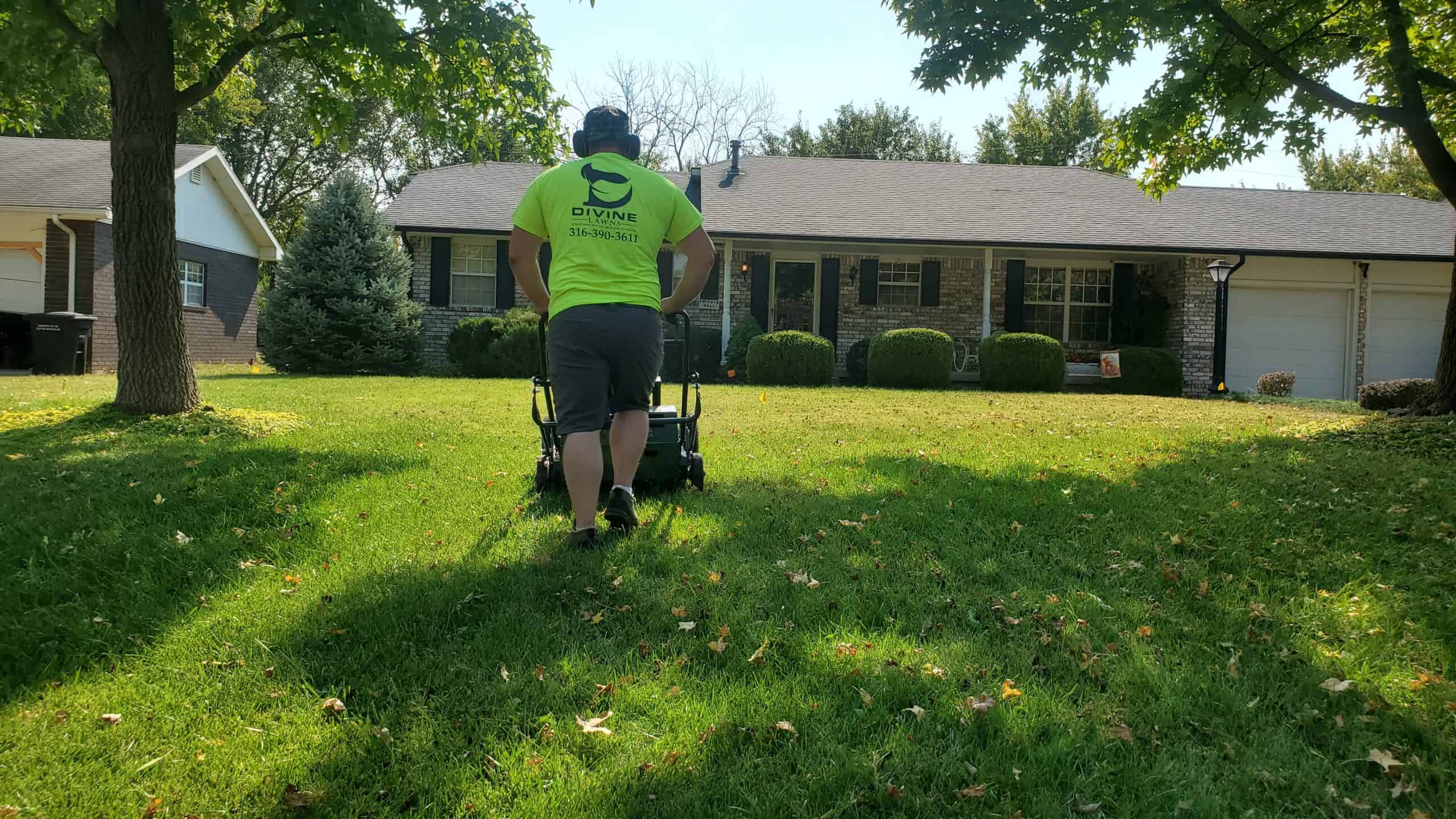 Worker in a neon shirt mowing a front lawn under tree shade, facing a brick house with trimmed bushes and a double garage.