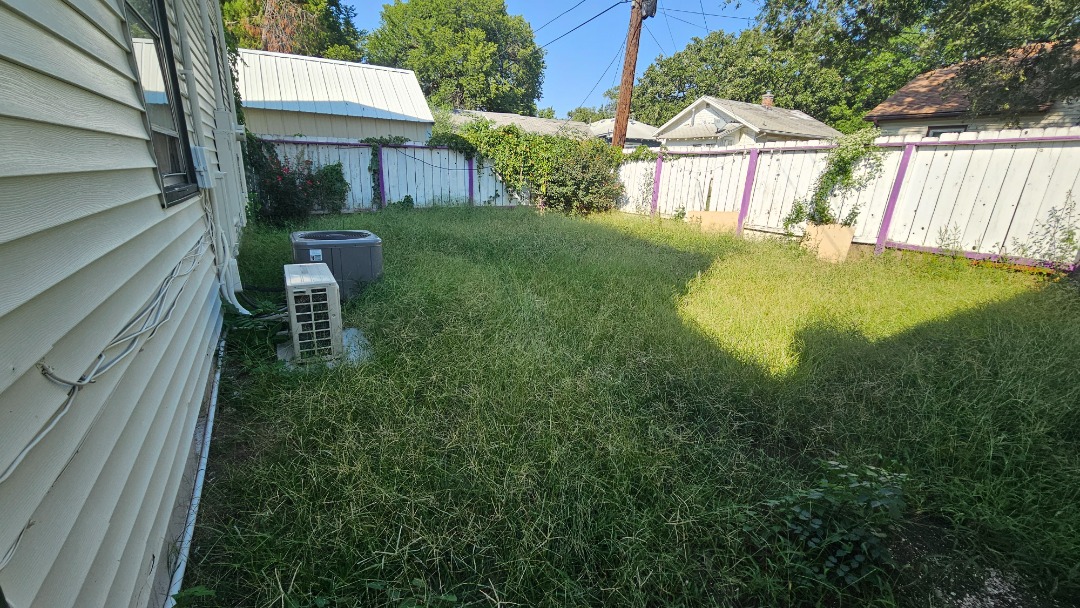 Overgrown backyard with tall grass, shaded areas, air conditioning unit, and white fence partially covered in vines and bushes.