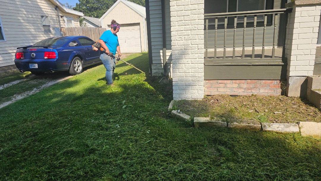 Landscaper in blue shirt using a string trimmer near a house foundation, next to a blue car and freshly cut green grass.