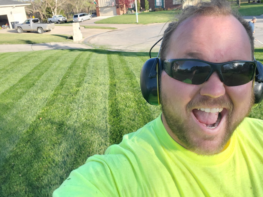 New landscaping hire takes selfie after mowing lawn, wearing ear protection and neon work shirt on residential property.