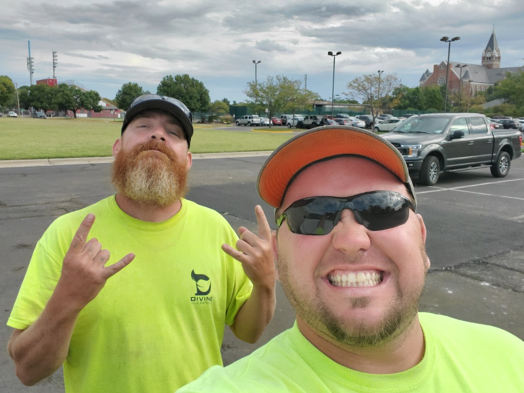 Two new landscaping hires pose in bright yellow shirts, smiling in a parking lot before heading to a job site with green space.